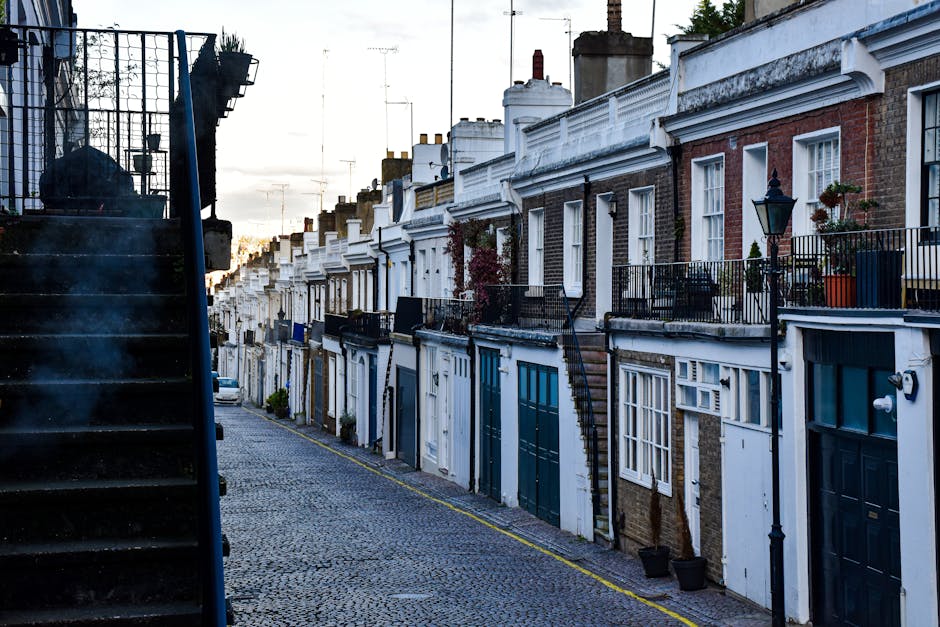 A row of terraced houses in Islington featuring brick and white-painted facades, with variously styled doors and windows, some with flower pots and hanging lanterns on small balconies. The street is paved with cobblestones, and a black metal staircase is visible on the left side, leading up to a building entrance. Several houses have external metal staircases and small garden areas with potted plants. The scene is captured during daylight, possibly in the late afternoon, with soft natural lighting illuminating the street. This environment depicts a typical residential area suitable for home relocation and furniture transport, reflecting the contextual backdrop for professional removals services such as those provided by Man and Van Islington.