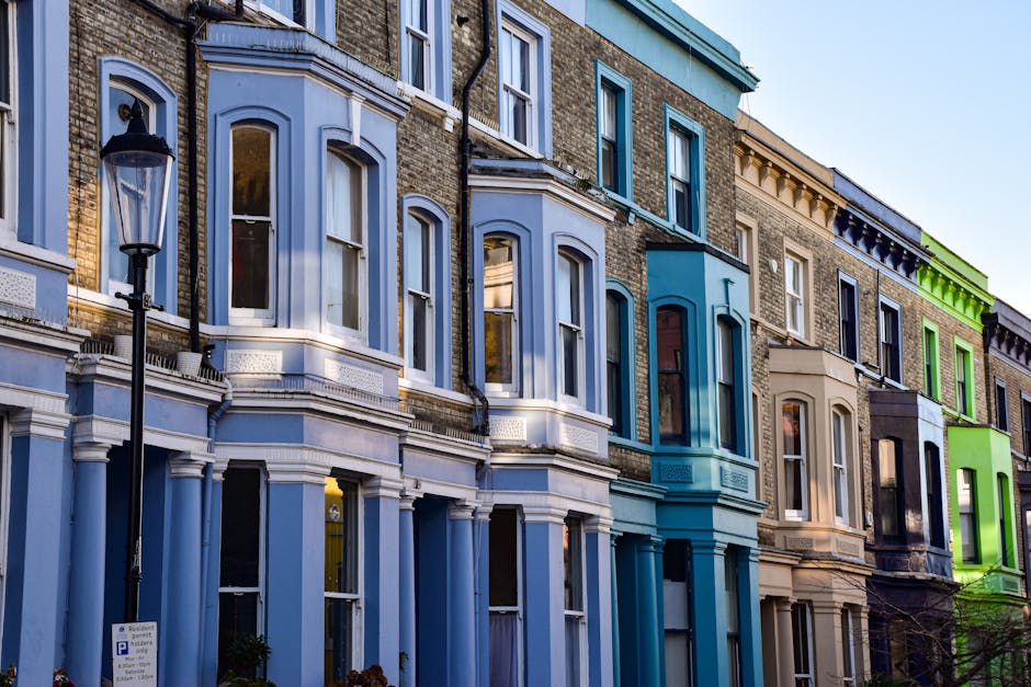 A wide view of a residential street in Islington with white Victorian-style terraced houses on both sides. The photograph captures a loading area where a man is seen preparing to load or unload furniture and boxes, which are partially visible on the pavement near a white van. Several cardboard boxes, some wrapped in plastic, are stacked and ready for moving, indicating a home relocation process. A few large pieces of wooden furniture, possibly wardrobes or cabinets, are being handled with the assistance of moving equipment such as trolleys or straps. The street is lined with parked cars and a black van, with trees featuring green and brown foliage providing a natural canopy overhead. Traffic lights at the intersection show red, and a streetlamp is visible in the background under an overcast sky. The scene exudes a professional and organized moving process, typical of house removals services like those offered by Man and Van Islington, highlighting the logistical effort involved in packing, loading, and transporting household items during a local move.