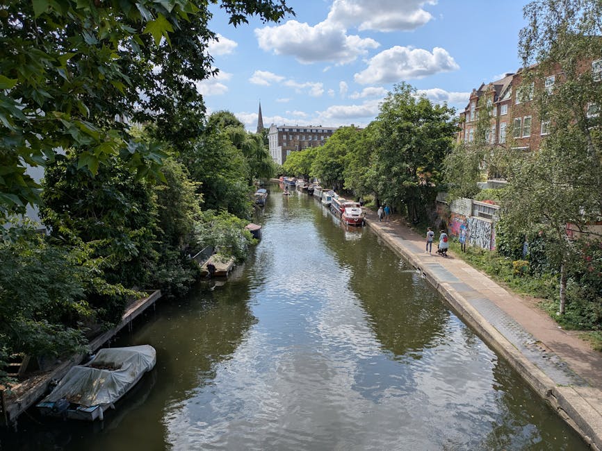 A daytime view of a narrow canal in a residential area, with calm water reflecting the blue sky and scattered clouds above. The canal is flanked by lush green trees on both sides, some extending over the water's edge. On the right side, a paved footpath runs parallel to the canal, where a few pedestrians are walking. There are several parked cars along the footpath, next to a row of brick buildings with large windows and decorative facades, typical of London architecture. On the left, boats are moored along the canal bank, one covered with a grey tarp. The scene appears peaceful and is indicative of a quiet, scenic urban environment suitable for home relocation projects involving furniture and belongings moving through outdoor surroundings. The setting aligns with services provided by Man and Van Islington for house removals and furniture transport, emphasizing the logistical aspect of loading or unloading near the waterway under natural daylight.