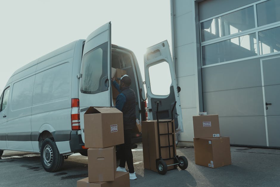 A professional mover from Man and Van Islington is loading cardboard boxes and packing materials into a white cargo van parked outside a building with large glass windows. The mover, wearing a dark uniform with a cap, is inside the van, arranging the boxes on a shelf or into the vehicle. Several additional boxes are placed on the pavement nearby, some with printed labels. A hand truck or dolly is situated next to a box on the ground, ready for transporting furniture or packed items. The scene takes place in bright daylight with the sun partially visible behind the van, indicating the home relocation process involves careful packing and loading of belongings into the vehicle, with attention to safety and organisation. The setting suggests a smooth furniture transport operation as part of a professional removals service for a house move or storage transfer.