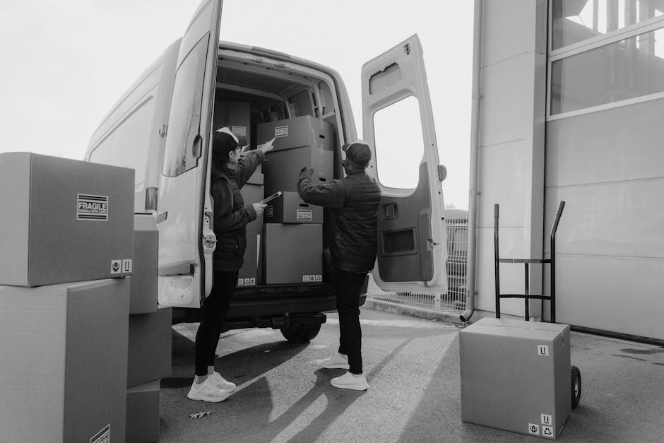 Two movers from Man and Van Islington are engaged in a home relocation process, unloading packed boxes and furniture from a white moving van parked outside a building with a driveway. The female mover, wearing a dark jacket and white sneakers, is inside the van passing a cardboard box or small appliance to the male mover, who is dressed in a dark jacket, dark trousers, and white shoes, reaching up to receive it. The open rear doors of the van reveal several packed items, including large cardboard boxes and wrapped furniture. Surrounding the van, there are additional cardboard boxes stacked nearby, some marked with fragile labels, ready for transport or storage. The environment appears to be an outdoor loading area with a paved surface, and a metal fence is visible in the background near a modern building with large windows. The photograph captures the logistics involved in furniture transport, packing, and loading, illustrating the careful handling of household items during a professional moving service provided by [COMPANY_NAME].