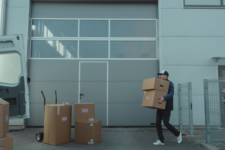 A professional mover from Man and Van Islington is loading cardboard boxes and packing materials into a white cargo van parked outside a building with large glass windows. The mover, wearing a dark uniform with a cap, is inside the van, arranging the boxes on a shelf or into the vehicle. Several additional boxes are placed on the pavement nearby, some with printed labels. A hand truck or dolly is situated next to a box on the ground, ready for transporting furniture or packed items. The scene takes place in bright daylight with the sun partially visible behind the van, indicating the home relocation process involves careful packing and loading of belongings into the vehicle, with attention to safety and organisation. The setting suggests a smooth furniture transport operation as part of a professional removals service for a house move or storage transfer.