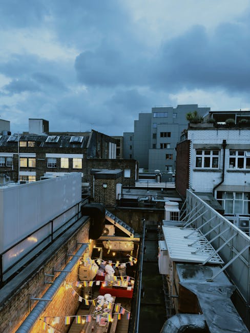 The image shows a view from a rooftop or balcony area adjacent to urban residential buildings, with a clear sky overhead. In the foreground, there is a narrow outdoor space with various objects related to packing and moving, including several cardboard boxes, some stacked and others scattered on a surface, likely prepared for a home or office relocation. A portable trolley or hand truck with a partially loaded box is visible, indicating ongoing efforts to transport items. The area is illuminated by warm artificial lighting, possibly string lights, creating a contrast with the natural daylight. Surrounding structures include a brick wall, metal railings, and industrial piping, characteristic of a city environment. In the background, multi-storey buildings with varied architectural styles are seen under a partly cloudy sky, suggesting a typical busy urban setting for house or office removals. This scene is connected to the services provided by Man and Van Islington, specialising in removals and furniture transport in the Islington area, supporting efficient packing, loading, and moving operations.