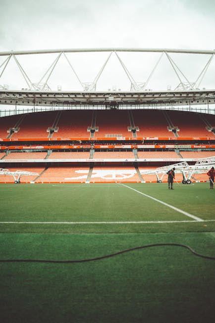 A view of a large football stadium with a green pitch in the foreground and orange seating sections in the background, partially covered by a white structural roof. Two movers, dressed in dark clothing, are visible on the field near a wheeled trolley loaded with packed boxes wrapped in plastic and carried on dollies, indicating a home relocation or furniture transport process. The scene is set during daytime with natural light illuminating the interior of the stadium, and the environment suggests a professional moving operation typical of house removals in London, such as those offered by Man and Van Islington, near Emirates Stadium and Holloway Road.