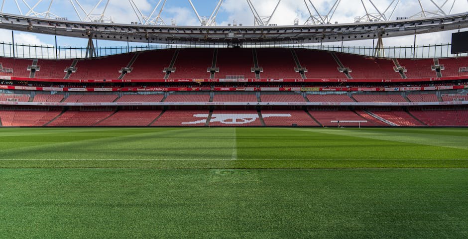 A wide view of an empty football stadium featuring a well-maintained grass pitch with white boundary and centre lines, surrounded by red stadium seating sections. The seating includes a large white logo or design in the middle of the stands. Above the stands, a modern roof structure with supporting beams extends over the seating area, under a partly cloudy sky. This setting showcases the arena typically used for football matches, with no visible players or spectators present, highlighting the venue's architecture and field condition related to sports and large event venues, which may be relevant for relocation or event setup services.