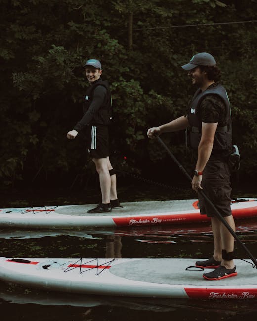 Two individuals standing on paddleboards in a waterway, surrounded by dense trees and foliage in the background. The person on the left, a woman, is smiling and looking towards the camera, wearing a black long-sleeve top, black shorts, a blue cap, and a life vest, while holding a paddle. The person on the right, a man, is looking down at the water, wearing a black t-shirt, black shorts, a grey cap, a black life vest, and holding a paddle, standing near the rear of the paddleboards. The boards are positioned on calm water with reflections of the trees above. The scene is set outdoors in natural light, capturing a moment of recreational water sport activity, which aligns with the theme of moving or outdoor adventure services provided by Man and Van Islington.