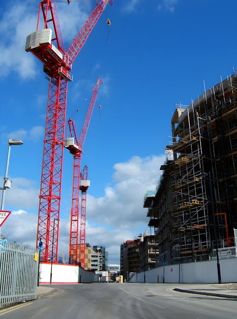 A construction site in Islington with two large red tower cranes towering against a bright blue sky with scattered white clouds. The cranes are positioned near a partially completed multi-storey building wrapped in scaffolding and temporary safety barriers, indicating active building work. The site is enclosed by a metal fence along the street, where a streetlamp and a sign are partially visible in the foreground. The scene captures the urban environment typical of city centre development projects, with the cranes used for transporting building materials to upper floors. The image reflects ongoing construction activity, which may involve logistics and planning in urban moving and site management, relevant to the context of house removals and moving services like those provided by Man and Van Islington.