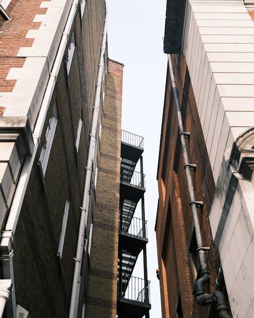 A narrow alleyway between two tall, multi-storey buildings during daytime, showing a metal fire escape attached to the brick wall on the left side, with several small landings and ladders. The buildin