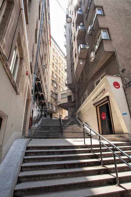 A narrow alleyway between two tall, multi-storey buildings during daytime, showing a metal fire escape attached to the brick wall on the left side, with several small landings and ladders. The buildin